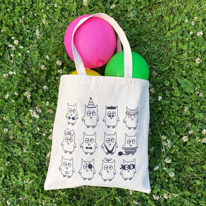 Natural canvas tote filled with bright balloons laying on grass and clover. The tote is printed in black with Drawful Owls. 